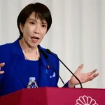 Takaichi, wearing a bright blue suit, gestures with her hands as she speaks at a podium