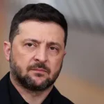 A close up image of Zelensky in front of a blurred European Union flag. He's wearing a black shirt and has a grim expression on