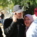 Raed wears a baseball cap. he's middle-aged with frey stubble. he's on his phone and has an arm around Ibtisam who is crying