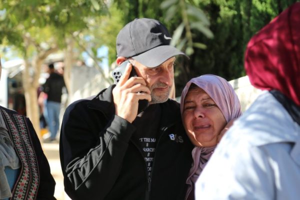Raed wears a baseball cap. he's middle-aged with frey stubble. he's on his phone and has an arm around Ibtisam who is crying