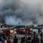 Onlookers gather as firefighters try to extinguish a fire that broke out in the cargo section of Hazrat Shahjalal International Airport in Dhaka on October 18, 2025. A large fire swept through the cargo terminal of Bangladesh's main international airport in Dhaka on October 18, forcing authorities to suspend all flights, officials said. (Photo by Maruf RAHMAN / AFP)