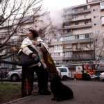 A woman stands with a dog next to the site of a damaged apartment building.